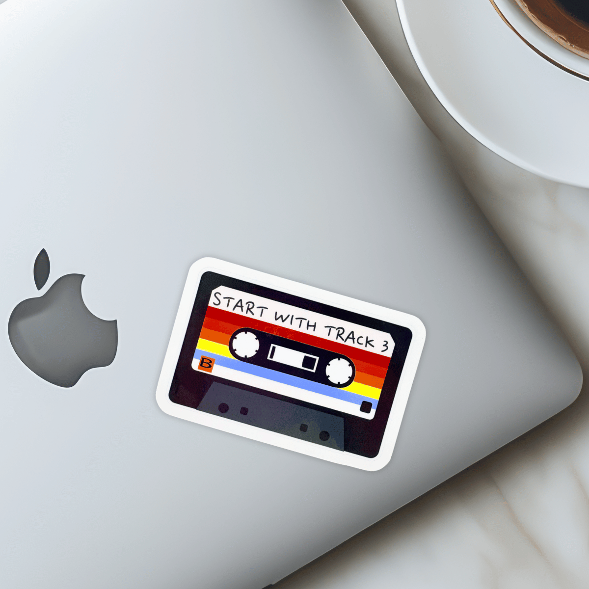 Sticker on a laptop with a rainbow cassette design and text 'Start with Track 3' next to a coffee cup.