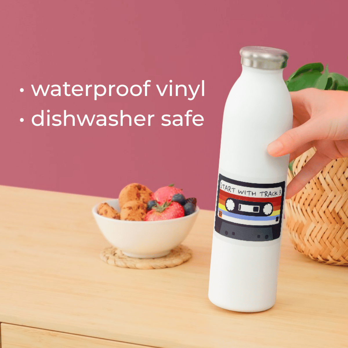 White water bottle with a cassette tape sticker on a wooden table with a bowl of fruit and cookies.