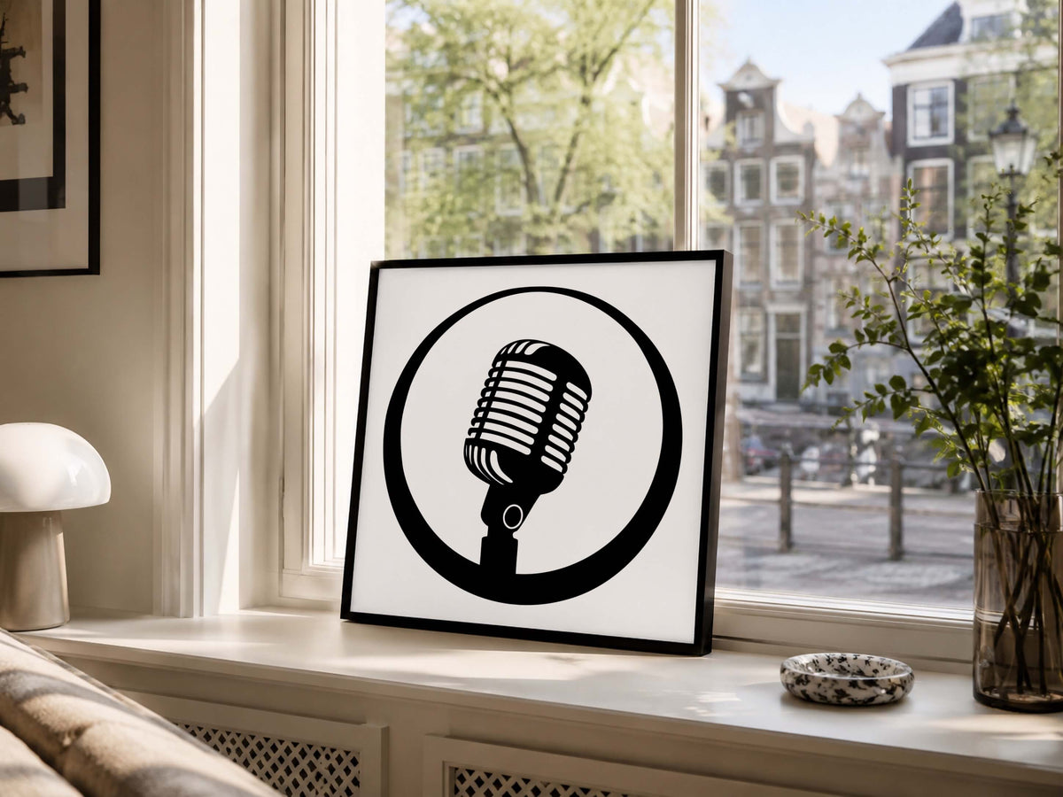 A square framed art print featuring a black retro microphone inside a black circle on a white background, leaning on a windowsill in a sunlit room with a city street and greenery visible outside.