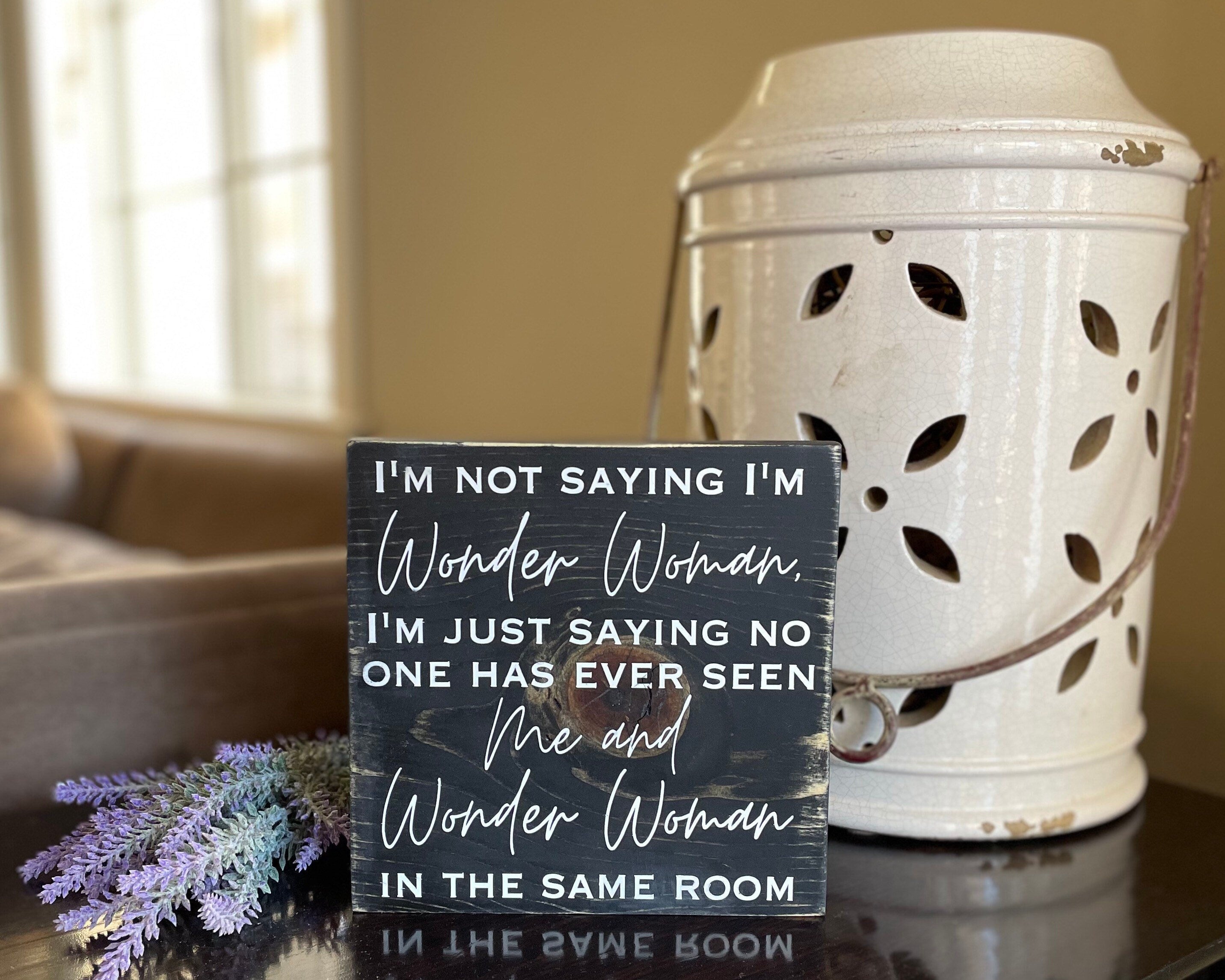medium sized square wood sign stained black with white lettering that reads, " I'm not saying I'm Wonder Woman, I'm just saying no one has ever seen me and Wonder Woman in the same room". All text is in all caps except for "Wonder Woman" and "Me and Wonder Woman". Text is centered, takes up the entire area and is on 7 lines. Glossy finish.