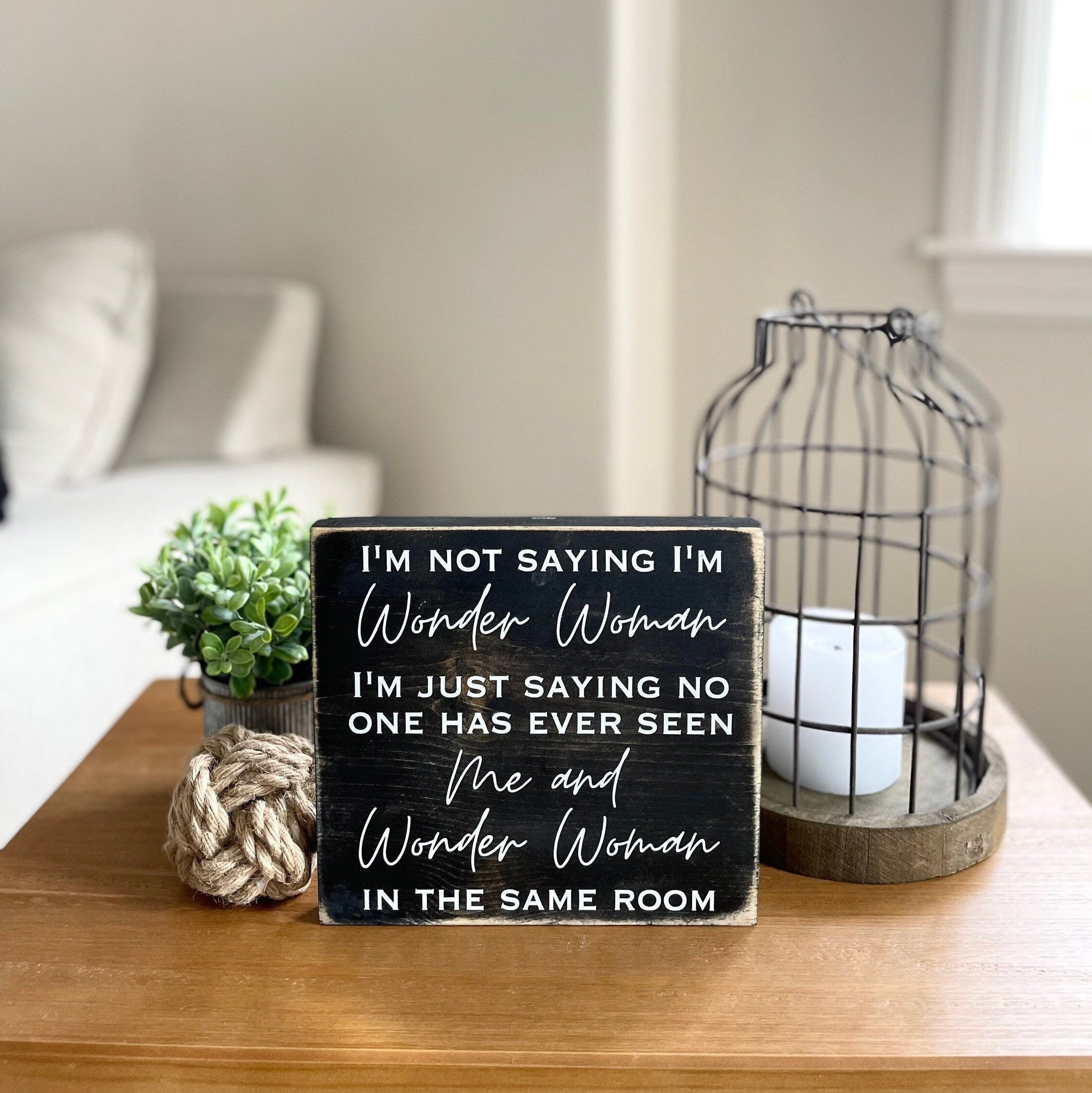 medium sized, square, rustic farmhouse style wood sign stained black with white lettering that reads, " I'm not saying I'm Wonder Woman, I'm just saying no one has ever seen me and Wonder Woman in the same room". All text is in all caps except for "Wonder Woman" and "Me and Wonder Woman" which is in a modern script font. Text is centered and takes up the entire area.