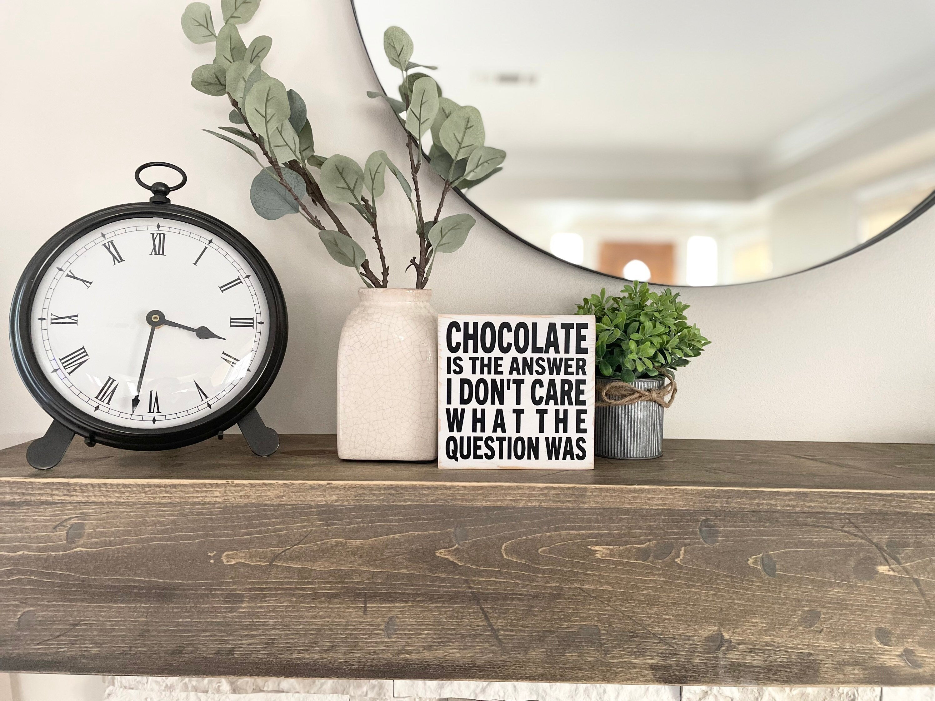 small, square, wood sign painted white with all caps text painted in black that reads: Chocolate is the answer I don't care what the question was. It is sitting on a wood mantle next to plants.