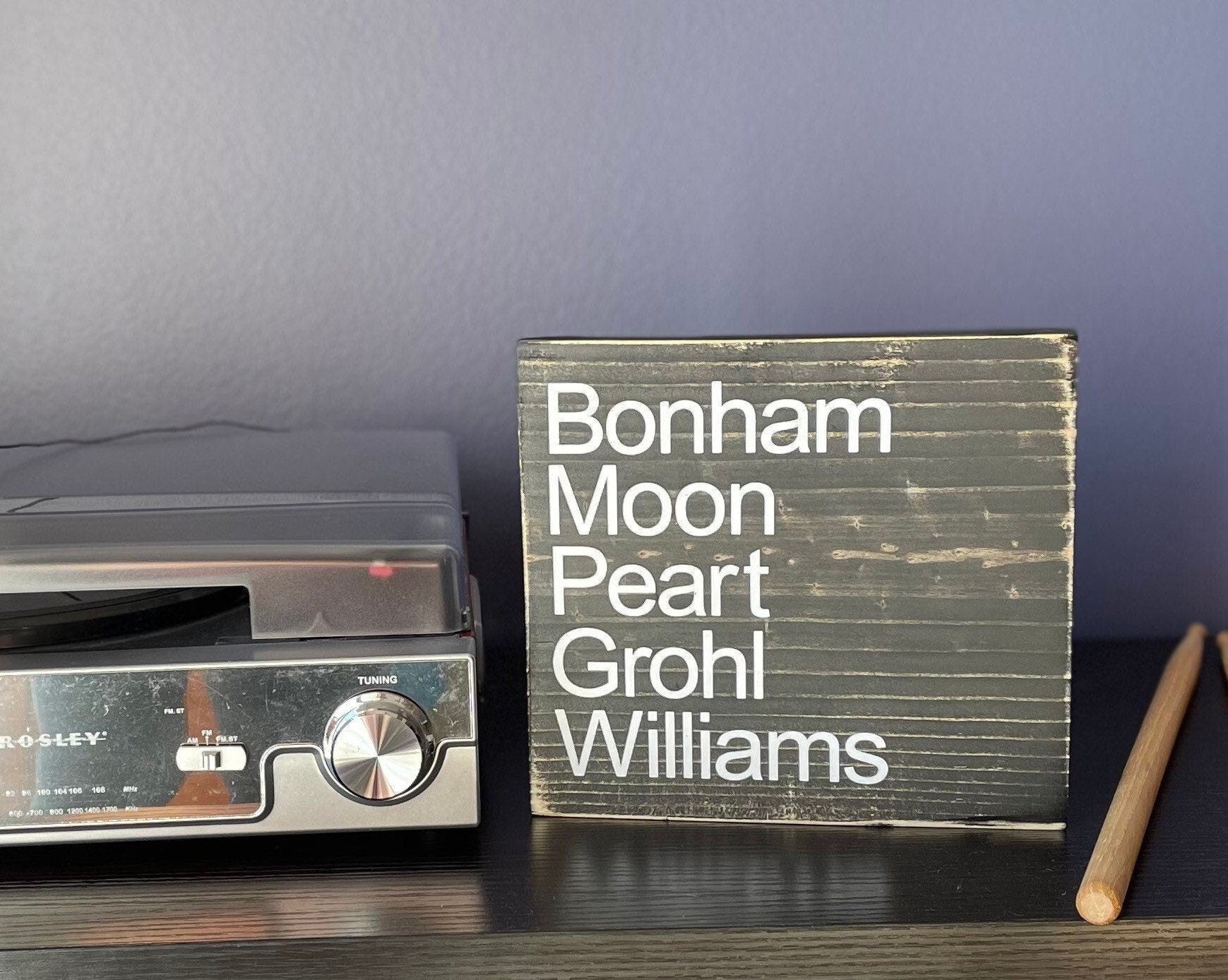 a square wood sign stained black with white, left justified, lettering that reads "Bonham Moon Peart Grohl Williams". T he names are listed top to bottom.