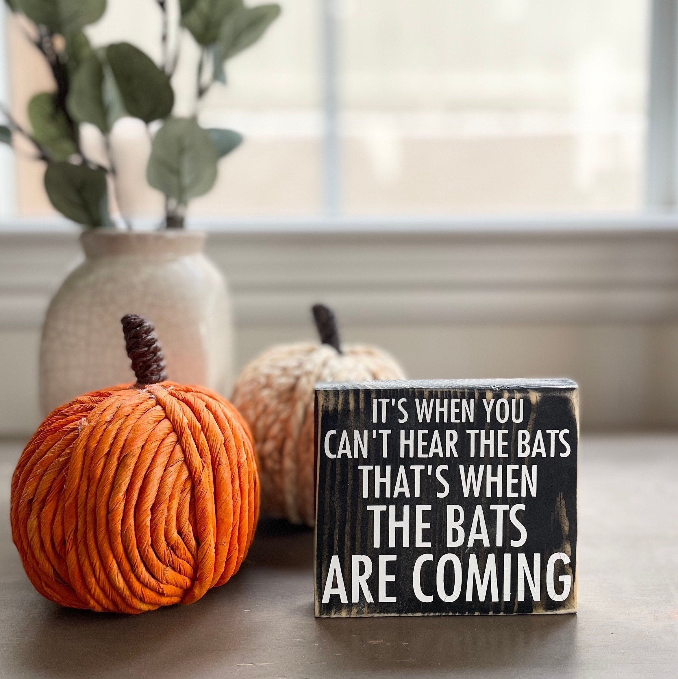 small square wood sign stained black with white lettering that reads in all caps "it's when you can't hear the bats that's when the bats are coming". The words are divided onto 5 lines. The size of the font gets bigger on each line