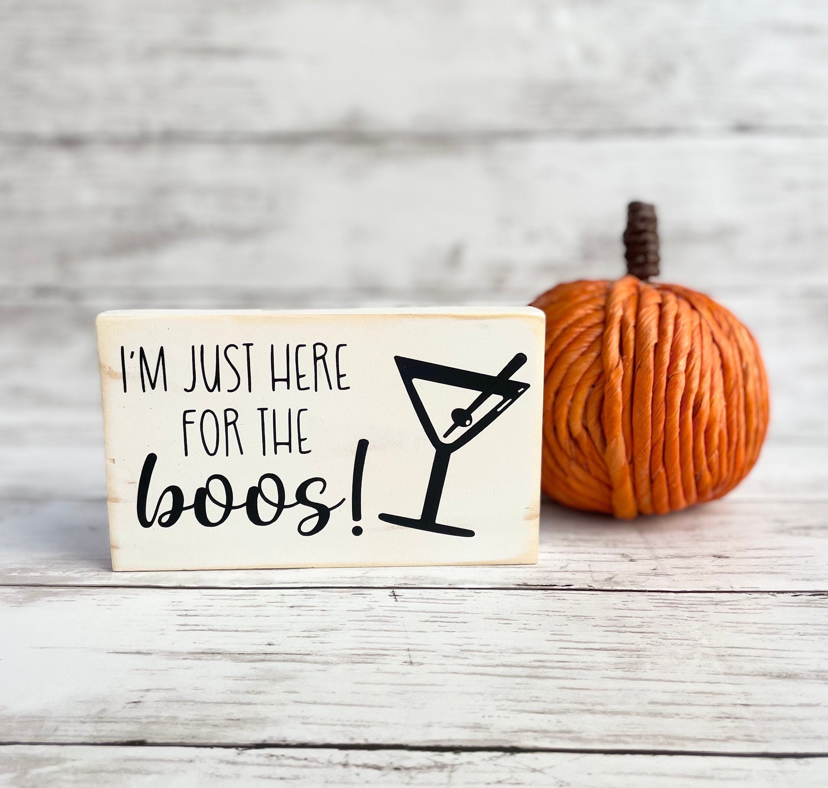 a small wood rectangular sign painted white with black lettering that reads "I'm just here for the boos!" All words are in all caps except for boos which is in a script font. The words are on the left side of the sign and an outline of a martini glass, with an olive on a toothpick, is on the right  