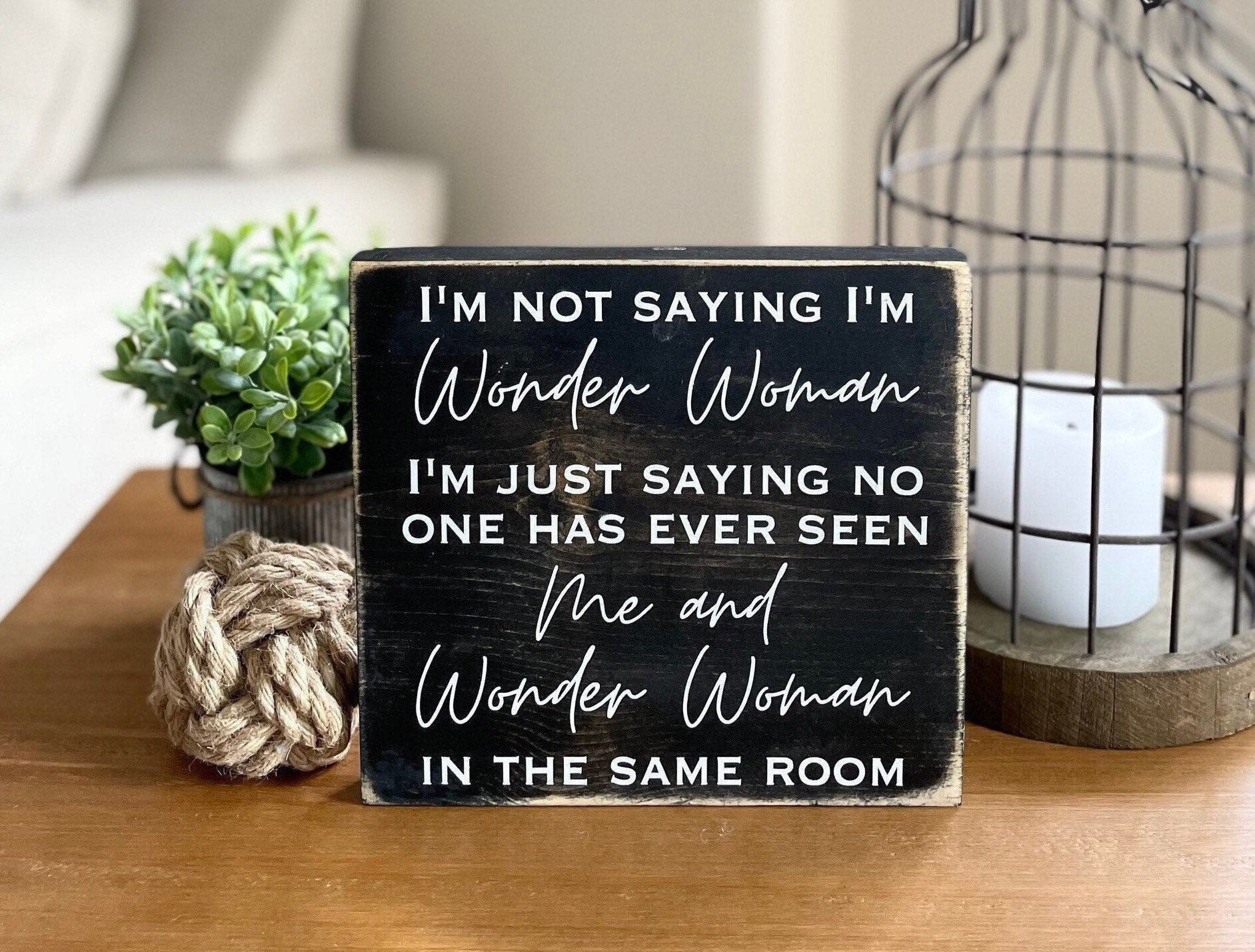 medium sized square wood sign stained black with white lettering that reads, " I'm not saying I'm Wonder Woman, I'm just saying no one has ever seen me and Wonder Woman in the same room". All text is in all caps except for "Wonder Woman" and "Me and Wonder Woman". Text is centered, takes up the entire area and is on 7 lines. Glossy finish.