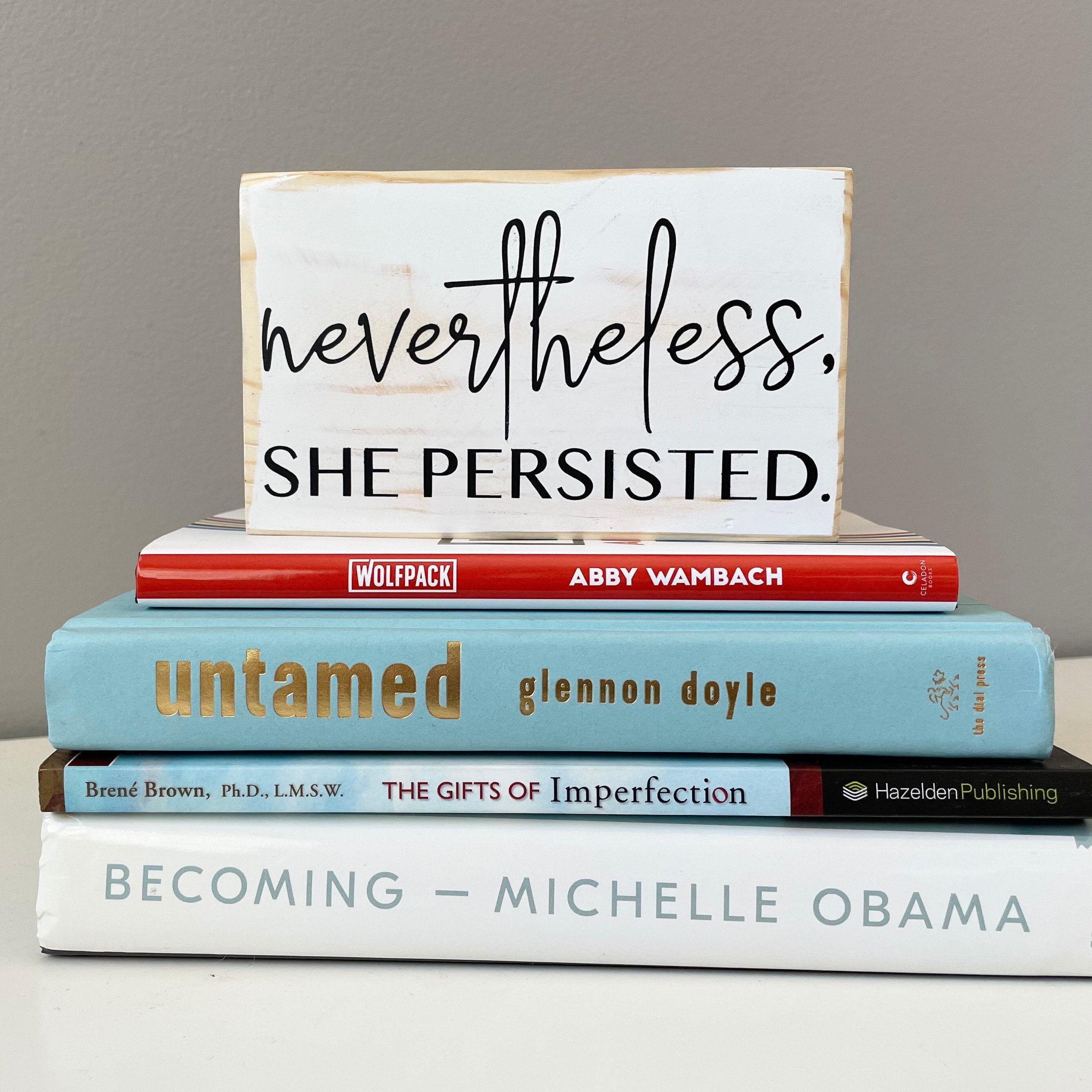 small rectangular wood sign painted white with black lettering that reads "Nevertheless, she persisted." Nevertheless is in script. She persisted is below it in all caps. 