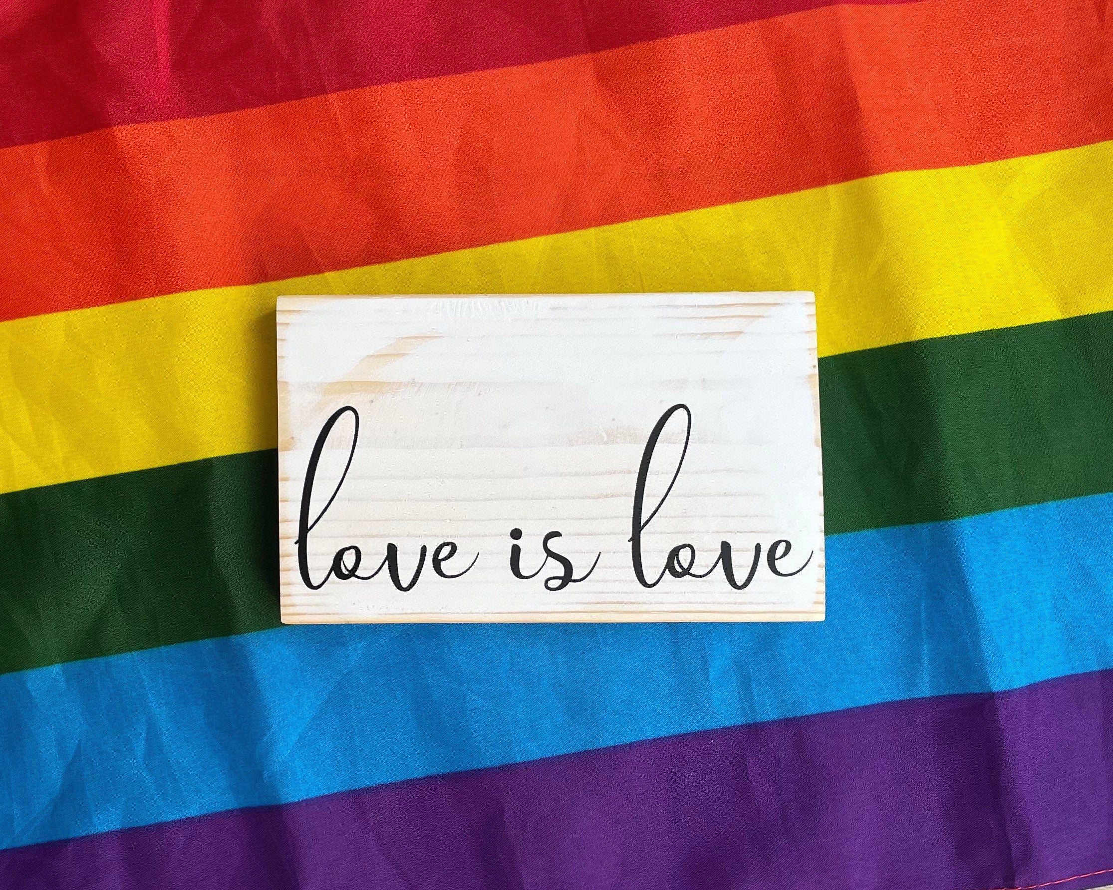 small wood rectangular sign painted white with black lettering that reads "love is love" in a delicate script font, all lowercase