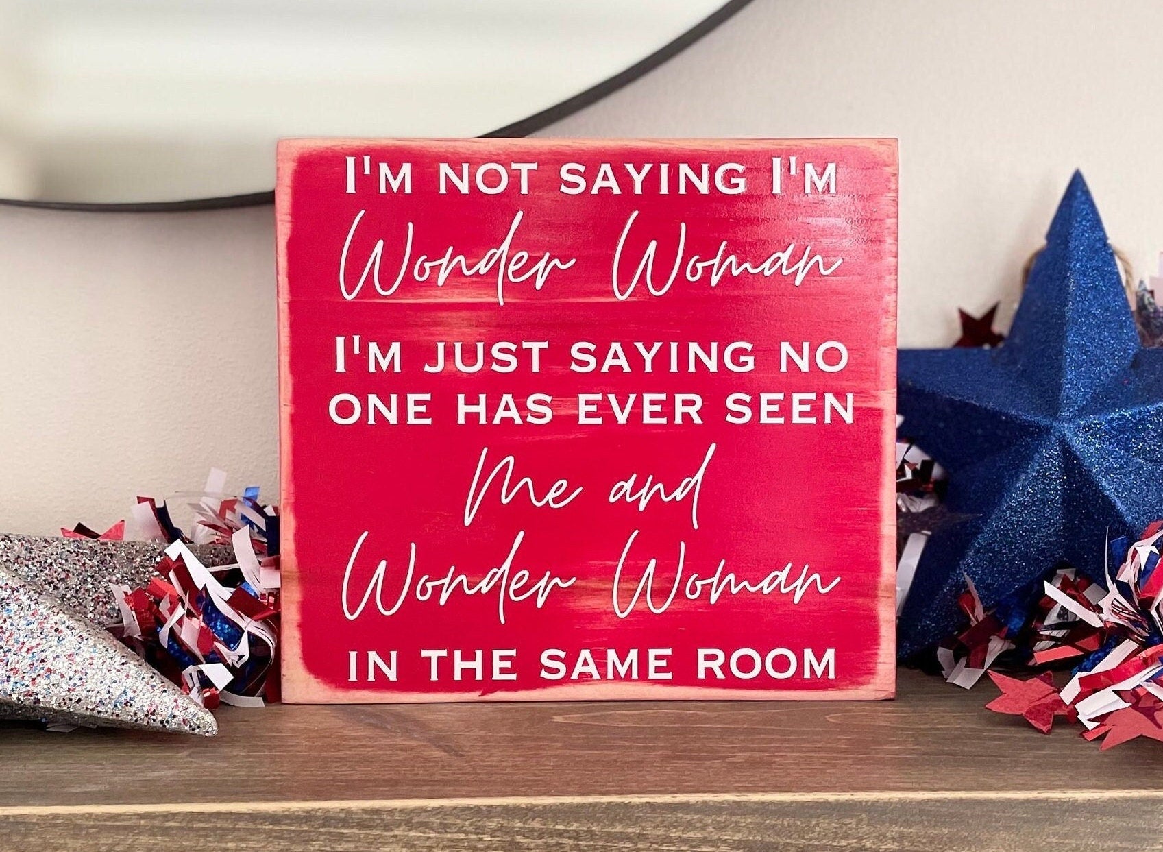 medium sized square wood sign painted red with white lettering that reads, " I'm not saying I'm Wonder Woman, I'm just saying no one has ever seen me and Wonder Woman in the same room". All text is in all caps except for "Wonder Woman" and "Me and Wonder Woman". Text is centered, takes up the entire area and is on 7 lines. Glossy finish.