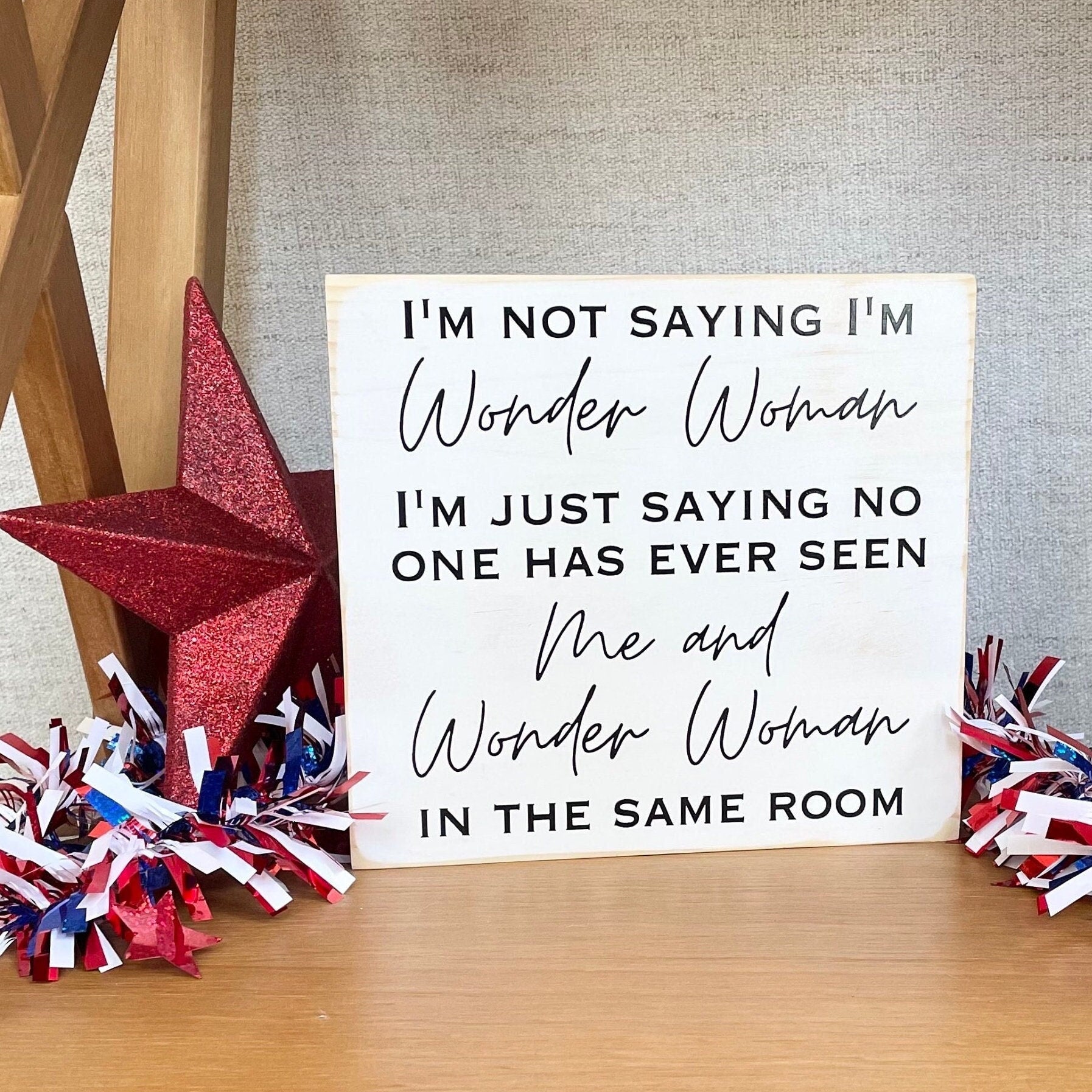 medium sized square wood sign painted white with black lettering that reads, " I'm not saying I'm Wonder Woman, I'm just saying no one has ever seen me and Wonder Woman in the same room". All text is in all caps except for "Wonder Woman" and "Me and Wonder Woman". Text is centered, takes up the entire area and is on 7 lines. Glossy finish.