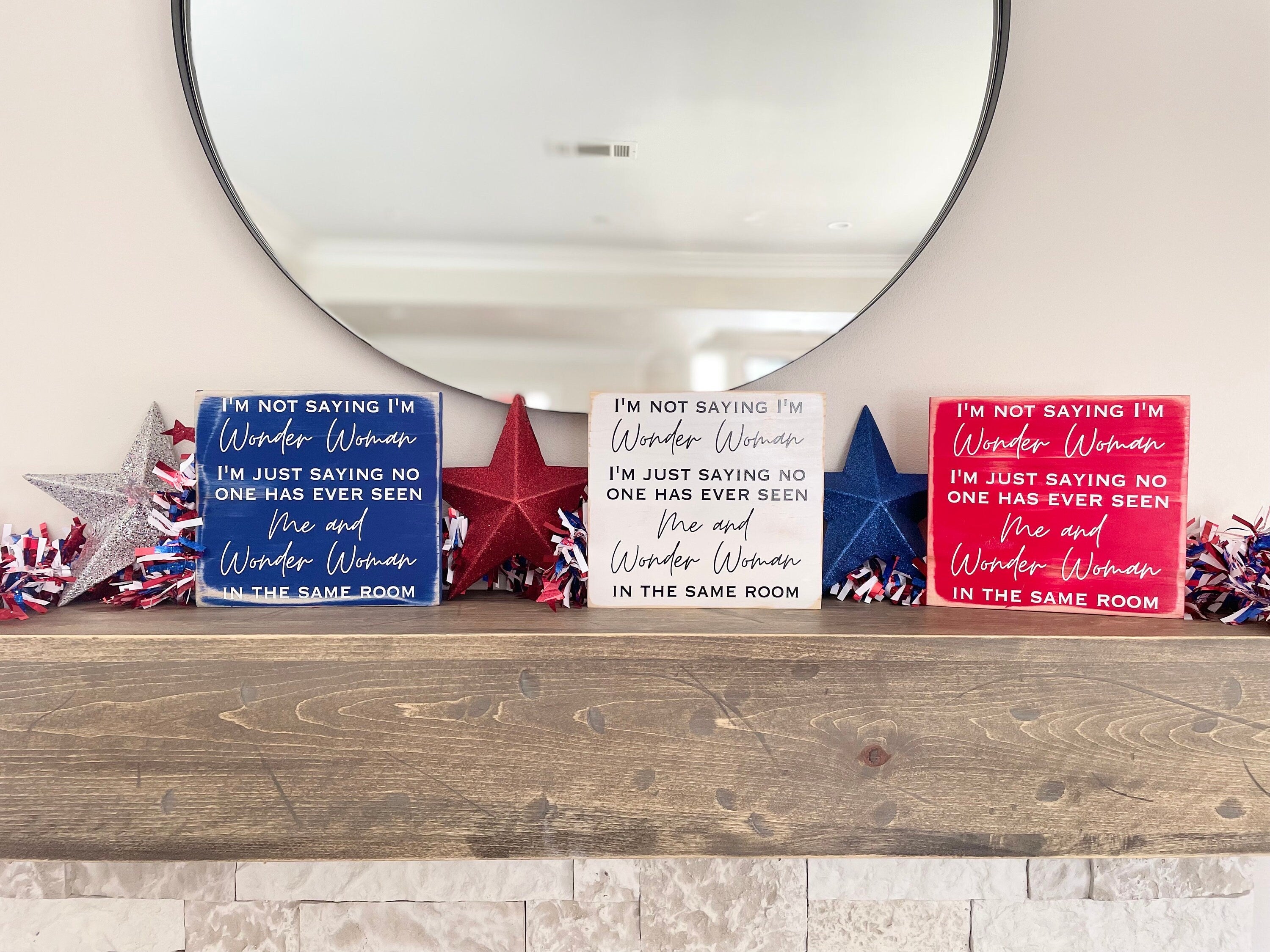 Three wonder woman signs on a mantle on a row each with the same quote on them. The one on the left is blue, the middle is white and the right is red. 