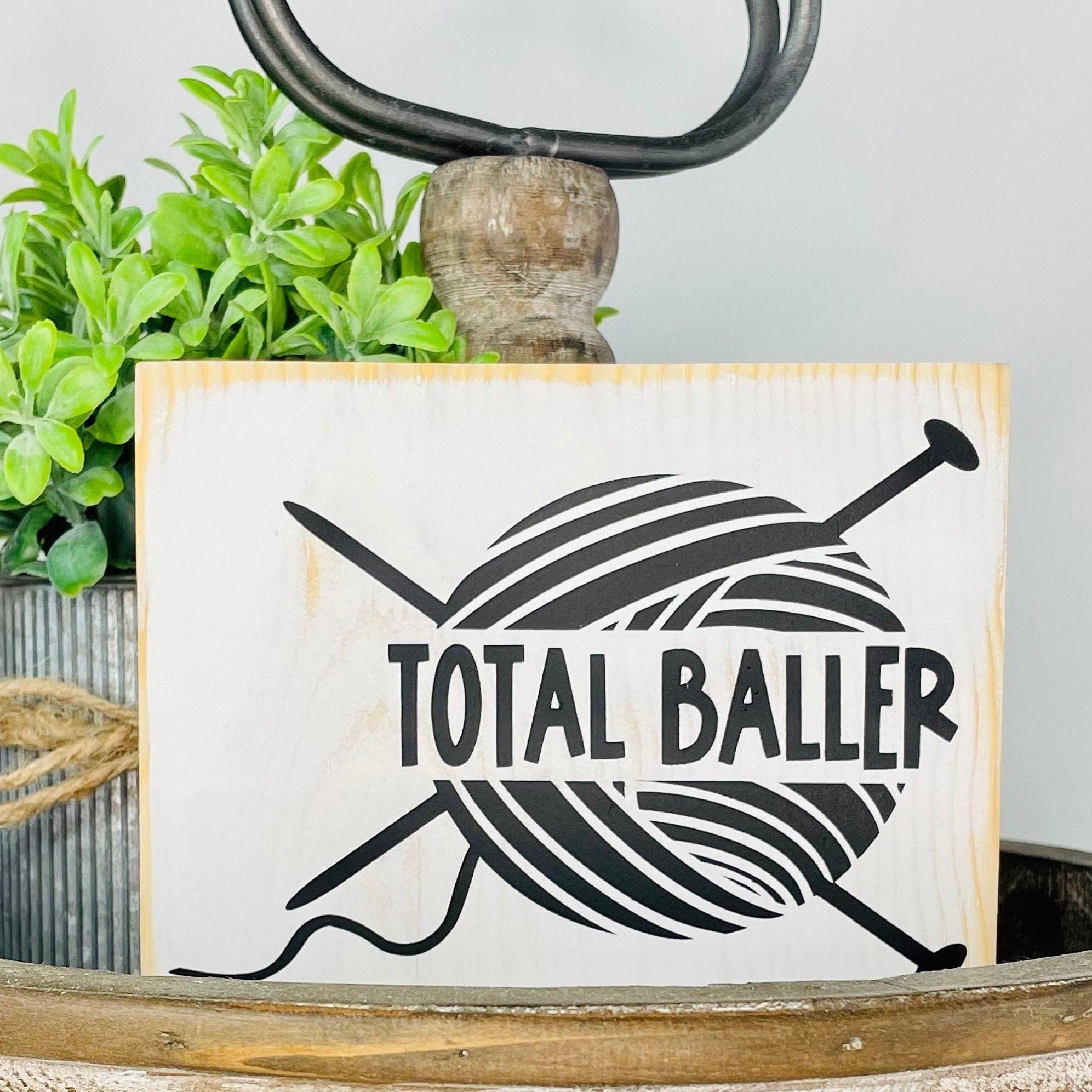 small rectangular wood sign painted white with an image of a ball of yarn with two knitting needles inserted into it making an X shape. The words "TOTAL BALLER" are written over the ball of yarn. The image and the text are in black.