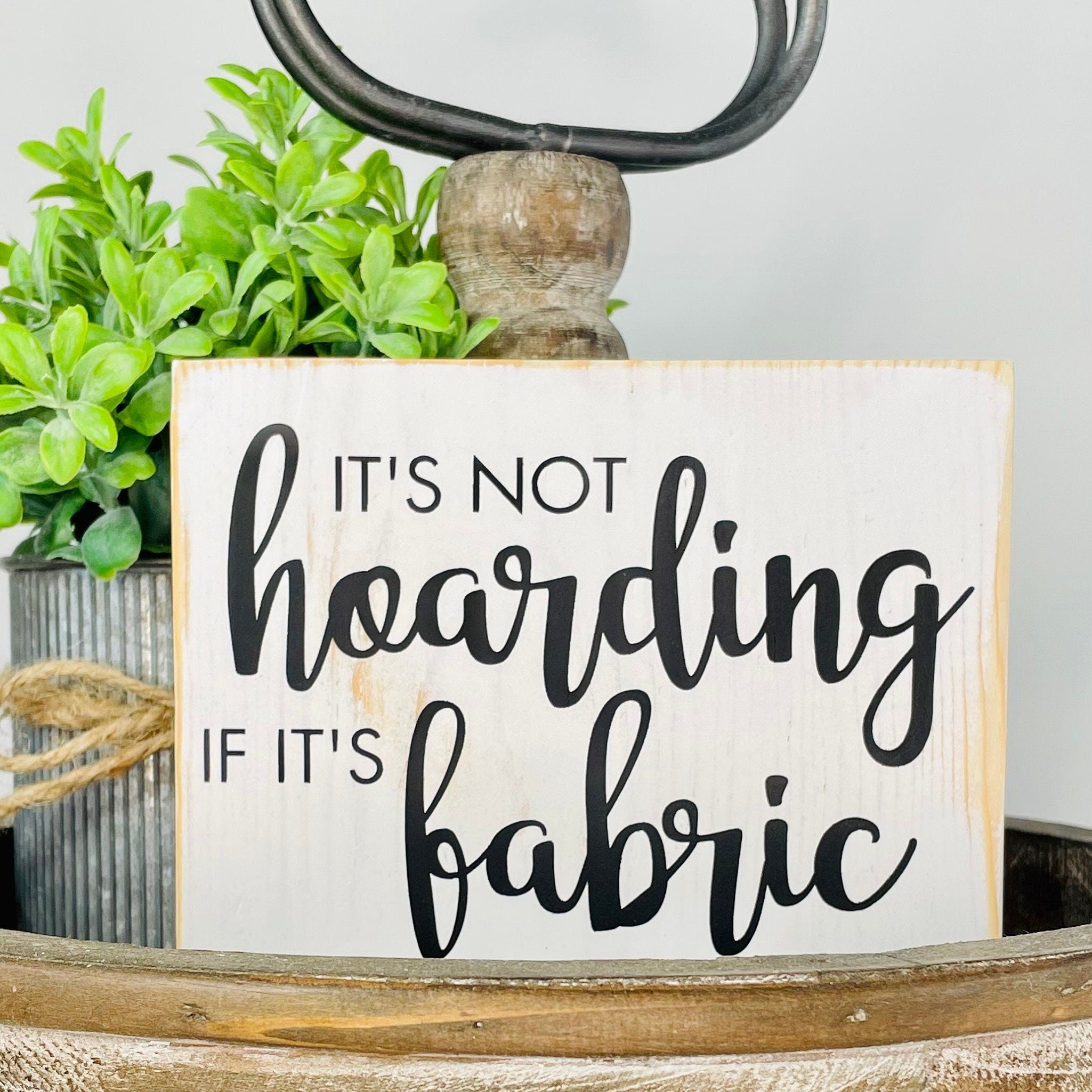 a small wood rectangular sign painted white with black lettering that reads "it's not hoarding if it's fabric". The words "hoarding" and "fabric" are in a large script font. The other words are in a thin font, all caps. 