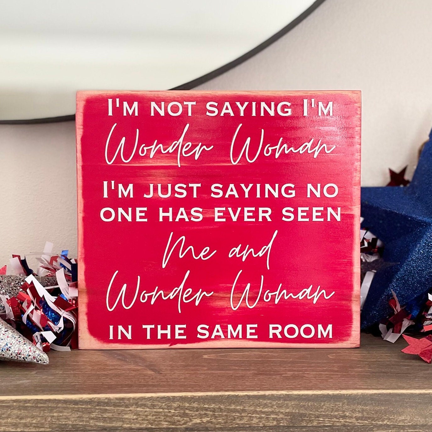 medium sized square wood sign painted red with white lettering that reads, " I'm not saying I'm Wonder Woman, I'm just saying no one has ever seen me and Wonder Woman in the same room". All text is in all caps except for "Wonder Woman" and "Me and Wonder Woman". Text is centered, takes up the entire area and is on 7 lines. Matte finish.