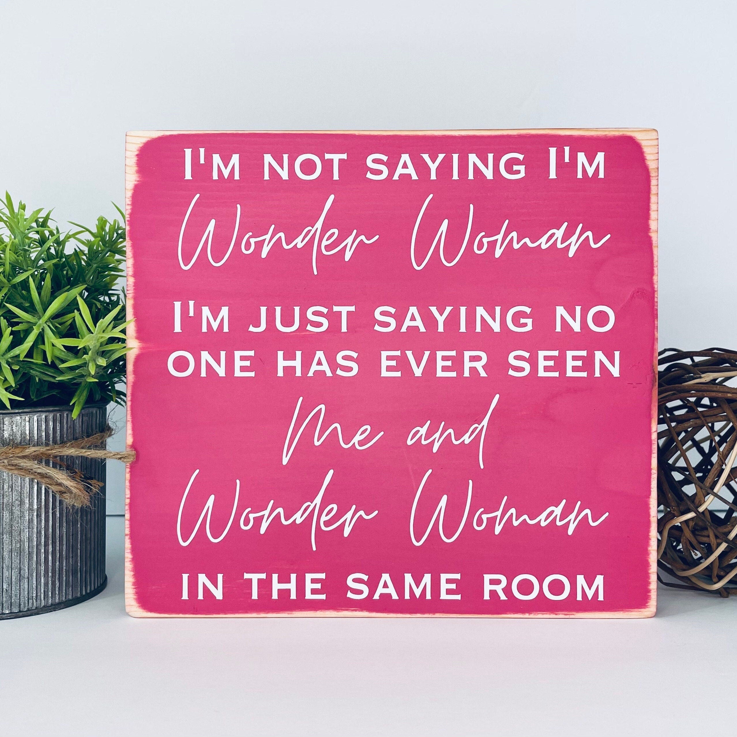 medium sized square wood sign painted pink with white lettering that reads, " I'm not saying I'm Wonder Woman, I'm just saying no one has ever seen me and Wonder Woman in the same room". All text is in all caps except for "Wonder Woman" and "Me and Wonder Woman". Text is centered, takes up the entire area and is on 7 lines. Matte finish.
