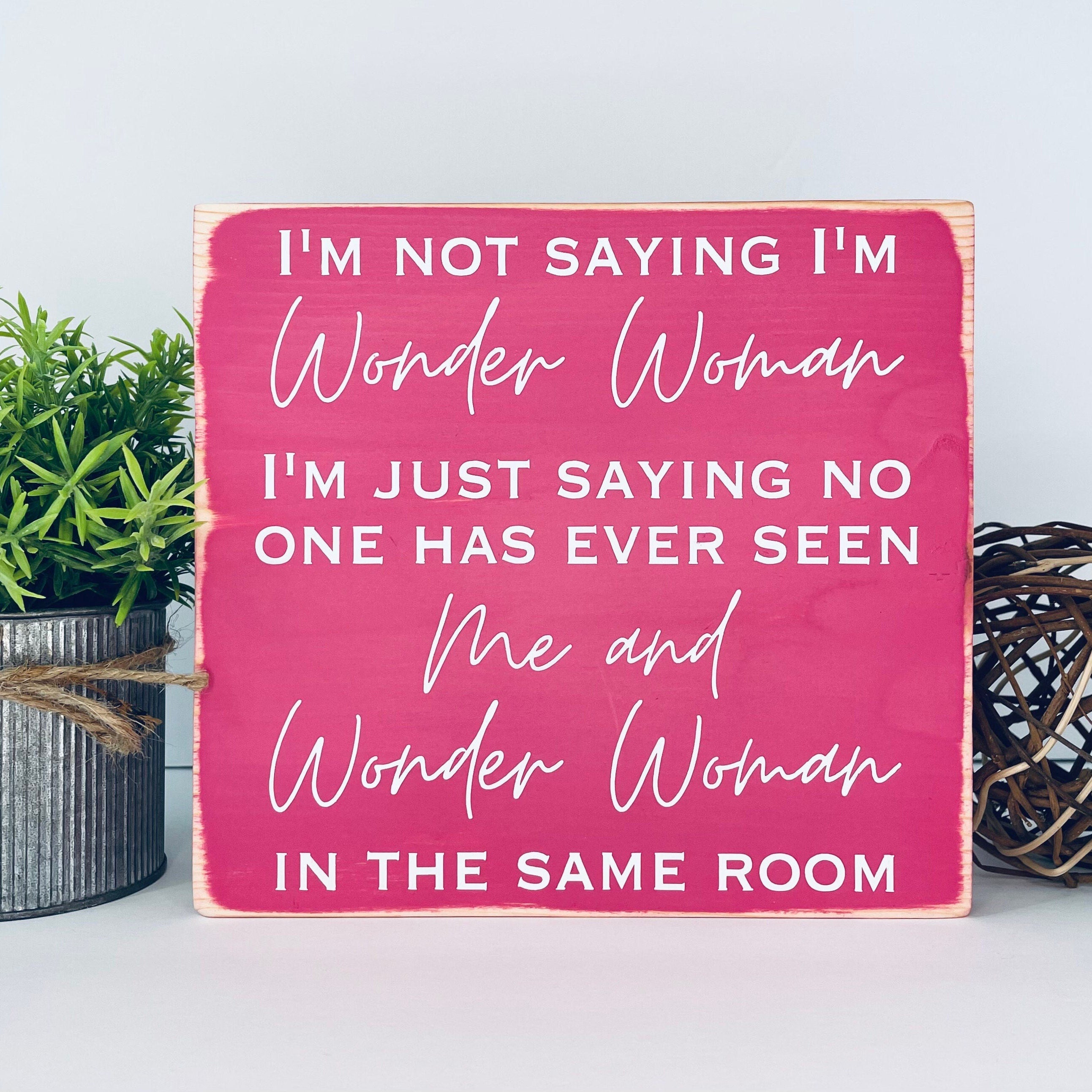 medium sized square wood sign painted pink with white lettering that reads, " I'm not saying I'm Wonder Woman, I'm just saying no one has ever seen me and Wonder Woman in the same room". All text is in all caps except for "Wonder Woman" and "Me and Wonder Woman". Text is centered, takes up the entire area and is on 7 lines. Matte finish.