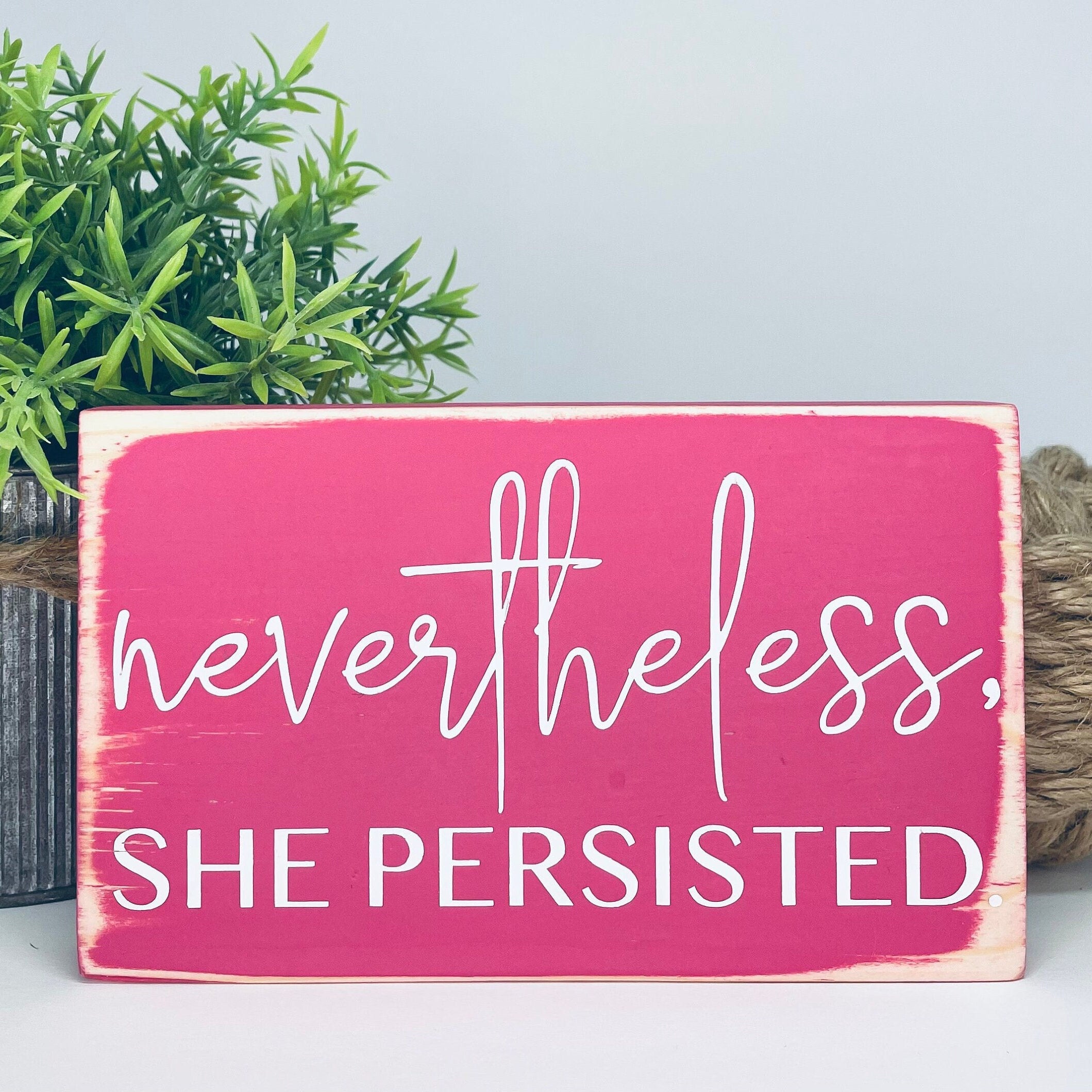 small rectangular wood sign painted hot pink with white lettering that reads "Nevertheless, she persisted." Nevertheless is in script. She persisted is below it in all caps.