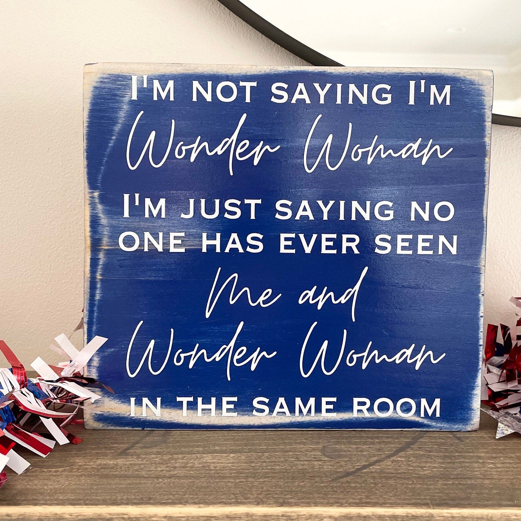 medium sized square wood sign painted blue with white lettering that reads, " I'm not saying I'm Wonder Woman, I'm just saying no one has ever seen me and Wonder Woman in the same room". All text is in all caps except for "Wonder Woman" and "Me and Wonder Woman". Text is centered, takes up the entire area and is on 7 lines. Matte finish.