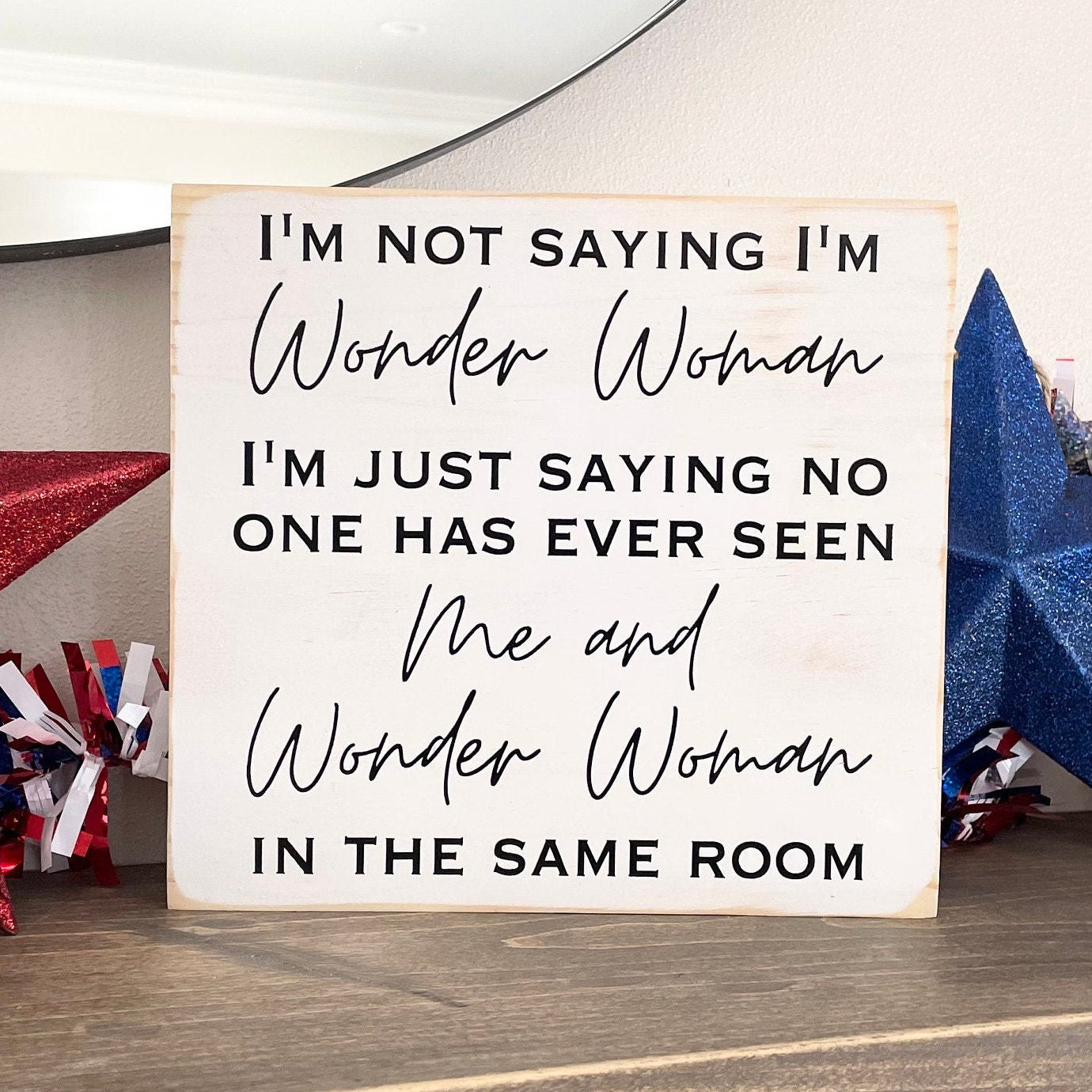 medium sized square wood sign painted white with black lettering that reads, " I'm not saying I'm Wonder Woman, I'm just saying no one has ever seen me and Wonder Woman in the same room". All text is in all caps except for "Wonder Woman" and "Me and Wonder Woman". Text is centered, takes up the entire area and is on 7 lines. Matte finish.