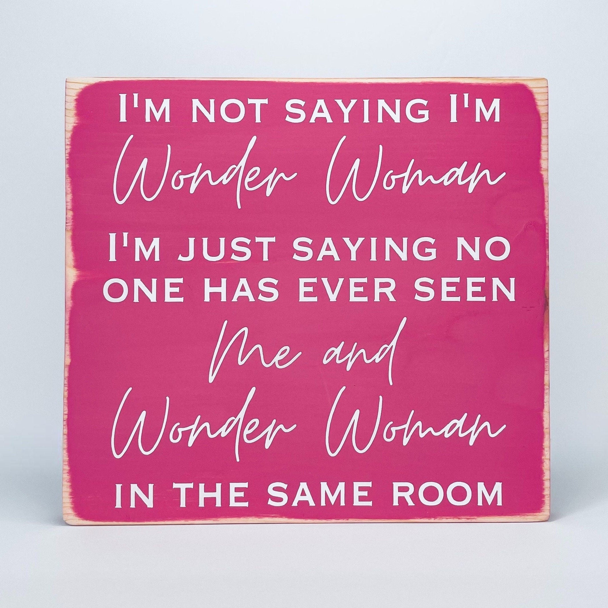 medium sized square wood sign painted pink with white lettering that reads, " I'm not saying I'm Wonder Woman, I'm just saying no one has ever seen me and Wonder Woman in the same room". All text is in all caps except for "Wonder Woman" and "Me and Wonder Woman". Text is centered, takes up the entire area and is on 7 lines. Matte finish.