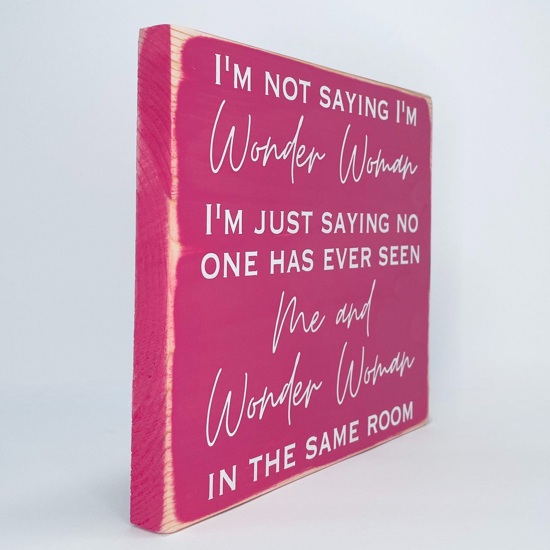 side view of a medium sized square wood sign painted pink with white lettering that reads, " I'm not saying I'm Wonder Woman, I'm just saying no one has ever seen me and Wonder Woman in the same room". All text is in all caps except for "Wonder Woman" and "Me and Wonder Woman". Text is centered, takes up the entire area and is on 7 lines. Matte finish.