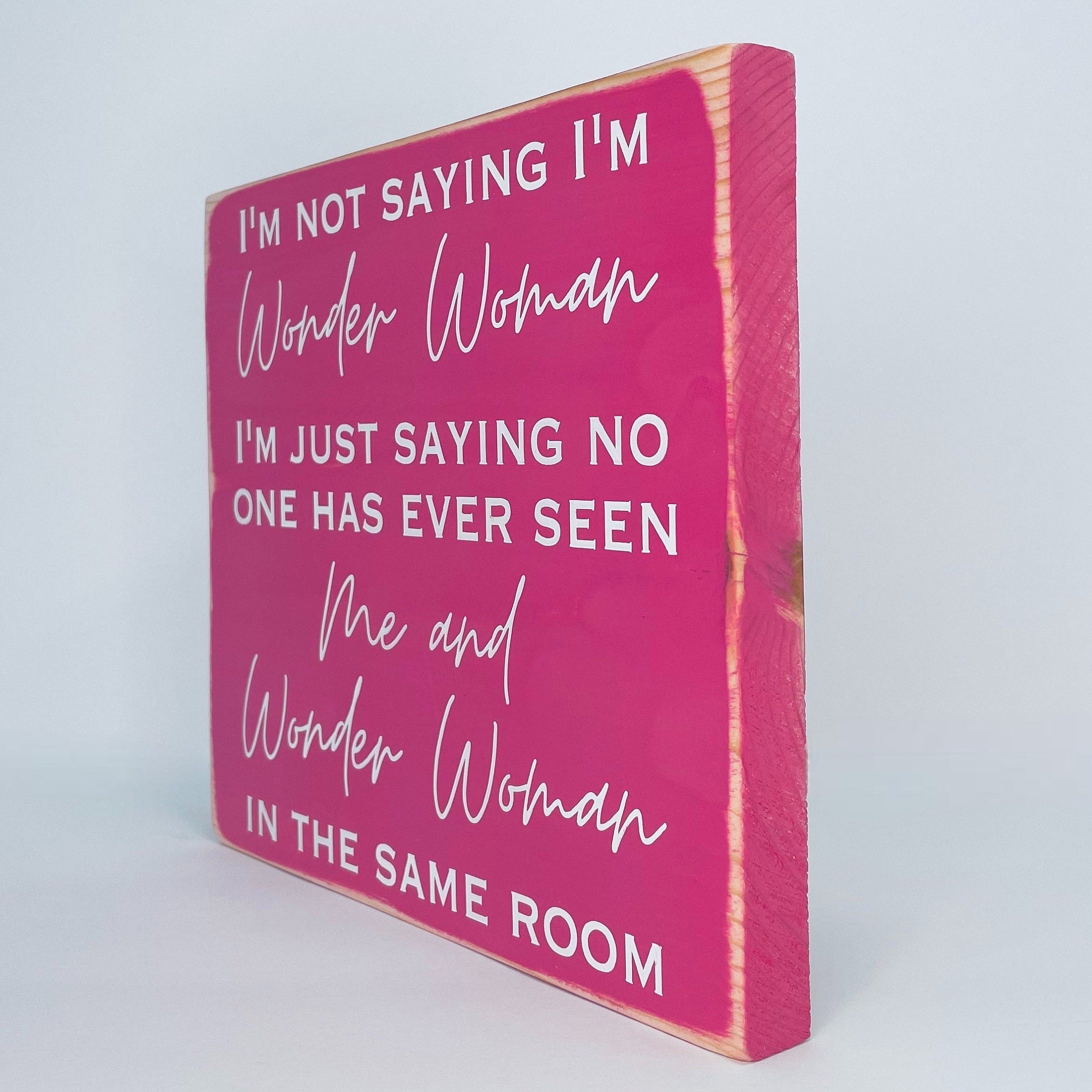 right side view of a medium sized square wood sign painted pink with white lettering that reads, " I'm not saying I'm Wonder Woman, I'm just saying no one has ever seen me and Wonder Woman in the same room". All text is in all caps except for "Wonder Woman" and "Me and Wonder Woman". Text is centered, takes up the entire area and is on 7 lines. Matte finish.